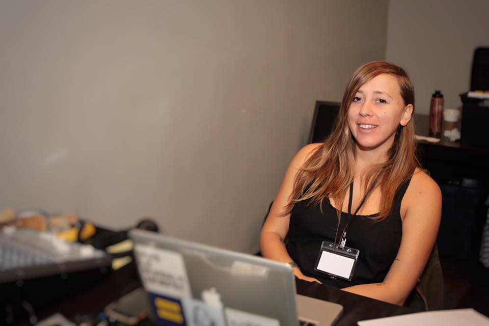 A warm, candid portrait of the Montes Media Collective founder seated behind a technical production desk. They have long, light-brown hair, are wearing a black tank top and an event lanyard with a badge, and smile directly at the camera with a welcoming expression. In the soft-focused foreground sits an open laptop decorated with stickers alongside an audio mixing console on the left side of the table. The background features a clean, neutral grey wall.