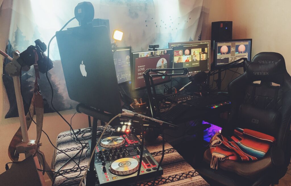 A high-end home production studio and digital broadcast workspace. In the foreground, a table covered in a neutral-toned striped textile holds a professional two-channel DJ controller alongside an elevated Apple laptop and a DSLR camera on a tripod. The main desk features a massive four-screen monitor array displaying a live Zoom video call gallery, a digital presentation slide that reads "WORKSHOP LOVE...", and media management interfaces. A professional microphone is mounted on an adjustable boom arm alongside warm LED panel lights. On the right, a black high-back gaming chair sits with a colorful, fringed, striped serape blanket draped over the seat. The background is softened by a hanging mountain landscape tapestry.