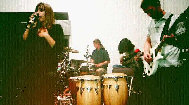 A nostalgic, grainy live band photograph from a stage performance. On the far left, the Montes Media Collective founder is singing passionately into a microphone, wearing a black long-sleeved shirt. In the foreground, a pair of wooden conga drums sits in front of a drum kit, where a drummer is playing in the background. On the right, a guitarist plays a white electric guitar next to a seated bass player, all performing together in a dimly lit, atmospheric rehearsal or venue space.