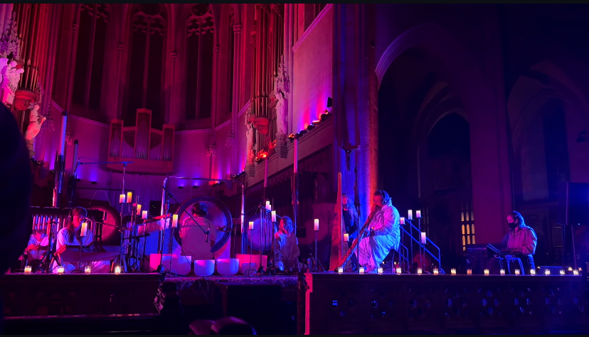 A wide stage shot of an ambient sound bath performance taking place on a grand cathedral altar, illuminated in deep magenta and purple theatrical lighting. The front of the stage is lined with dozens of glowing tea lights and tall candles. Several musicians are spaced across the stage playing a large metallic gong, crystal singing bowls, a harp, and a flute. On the far right side of the stage, the primary audio engineer (A1) is positioned at a technical station, actively managing the live sound under a wash of cool blue light.