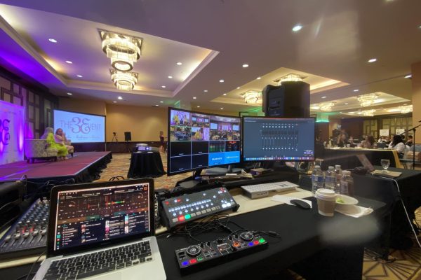 A technical director's perspective from the front-of-house control station at a live corporate event. In the foreground, a black production desk is meticulously organized with a laptop running digital audio software, a compact DJ controller, a video production switcher console, and dual large monitors. One monitor displays a multi-view video feed of the live stage and audience, and the other shows an interactive software audio mixer interface. In the background, a stage illuminated in vibrant magenta and purple features two speakers seated in armchairs. A large projection screen displays the branding for "THE 3E EVENT" inside a well-lit hotel ballroom.