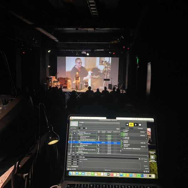 A technical director's perspective from the control station during Denice Frohman's one-woman show, "Esto No Tiene Nombre," at MACLA. In the sharp focus of the foreground, a laptop screen displays the QLab software cue list under the show file name "ESTOshowMACLAPerformance," with active cues visible for video projections like "glitch," "Ruthie Borie - Barbershop Tour," and "Mom Interview," alongside a small glowing desk lamp. In the midground, the dark silhouettes of a seated theater audience look toward the stage. On the stage, performer Denice Frohman stands in a black t-shirt and brown trousers under warm spotlights, speaking in front of a giant projection screen displaying a video of a Latina elder. The stage is dressed on the left with a cozy red velvet wingback armchair, lamps, and boxes, and a music stand on the right.