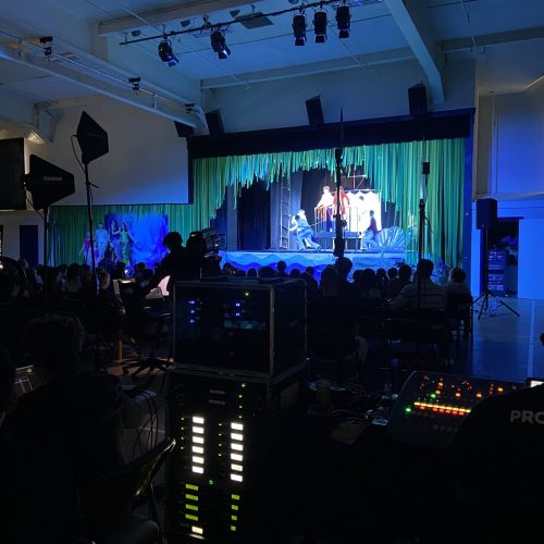 Behind-the-scenes view from the production booth of a live theatrical performance. In the darkened foreground, technical crew members sit at a control station, including one on the right wearing a black t-shirt that reads "PRODUCTION." The desk features an audio mixing console with glowing green and yellow indicator lights and stacks of professional AV rack equipment showing vertical light meters. In the background, a seated audience watches a brightly lit stage decorated with hanging green and blue streamers resembling an underwater scene, where actors are performing a live play under blue theatrical lighting, for a musical at a school called Hillbrook.