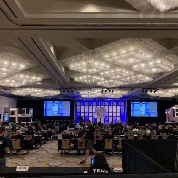 Behind-the-scenes view of a large-scale live event production inside a grand, brightly lit ballroom. In the foreground, a professional video camera sits on a tripod next to event crew members wearing black t-shirts with "TEAM" printed in bold white letters. In the background, a massive stage features three large projection screens lit in vibrant blue, displaying a live feed of a speaker, flanked by custom stage set designs. A large audience is seated at tables beneath a ceiling adorned with rows of glowing, geometric crystal chandeliers.