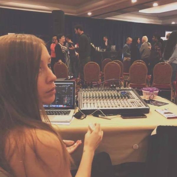 Behind-the-scenes photograph of the Montes Media Collective founder seated at a technical production table inside an event hall. In the foreground, an analog mixing board with multiple faders and knobs sits next to an open laptop displaying digital audio software. The founder, a human with long brown hair, looks out thoughtfully toward the room. In the soft-focused background, event attendees stand in small groups, talking and connecting near rows of empty conference chairs.