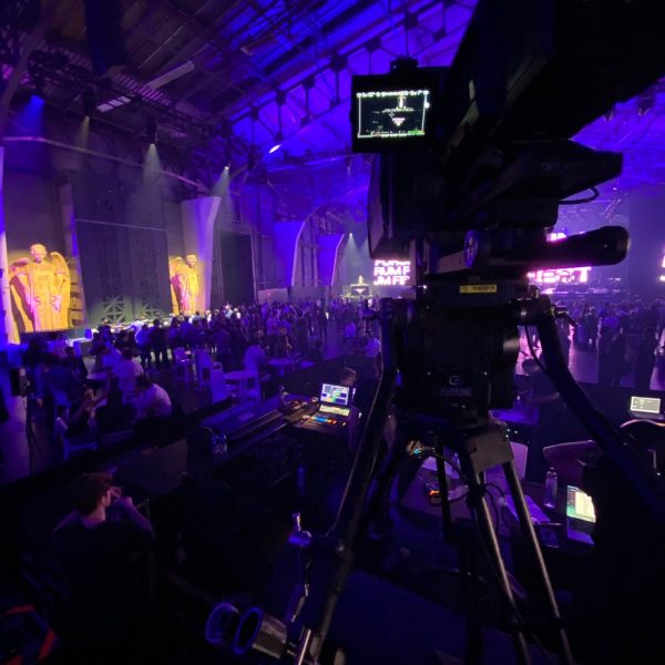 A high-angle, behind-the-scenes view from a raised technical production riser inside a massive, industrial warehouse-style event venue. In the sharp focus of the foreground, a large, professional broadcast video camera is mounted on a heavy-duty tripod, its small monitor screen illuminated. Below the platform, the technical booth is visible with crew members working at glowing laptops and a control console. The sprawling venue in the background is bathed in dramatic deep purple and blue lighting, filled with a large crowd of attendees. On the left wall, two giant, illuminated golden angel statues flank the space, while a bright stage screen glows in the far distance.