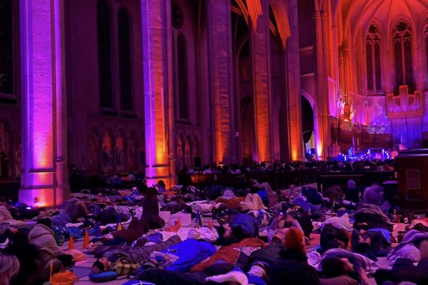 An immersive, large-scale community event taking place inside a grand gothic cathedral with soaring stone columns and high vaulted arches. The architectural pillars are dramatically illuminated in vibrant washes of magenta, purple, and warm orange up-lighting. On the stone floor below, a large crowd of people lies peacefully on yoga mats, tucked under blankets and sleeping bags, participating in a quiet sound bath or meditation journey.